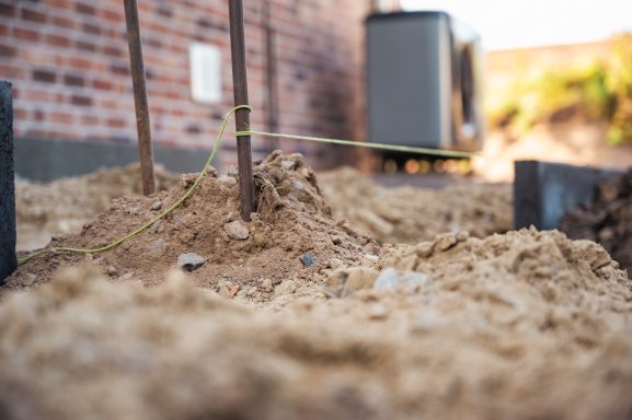 Baugrund mit Sand, erdige Fläche und einige Holzpfähle, im Hintergrund ein Gebäude.
