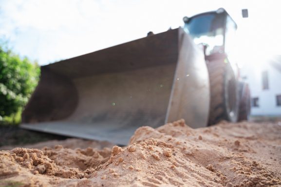 Bagger mit offenem Schaufel, Erde im Vordergrund, helle Himmelsszene.