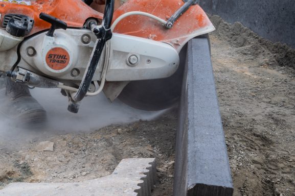 Sich in Bewegung befindliches Baugerät schneidet in Schotterboden. Dustwolken steigen auf.