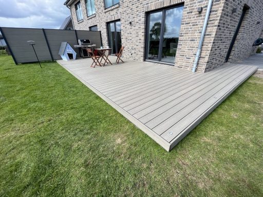 Holzterrasse mit Tisch und Stühlen vor einem modernen Haus, umgeben von Rasenfläche.