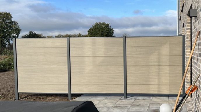 Zaun aus hellen Paneelen und grauen Pfosten, umgeben von einem Garten.