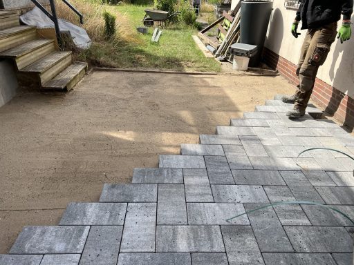 Fußweg mit grauen Pflastersteinen, daneben Treppen und eine unvollendete Fläche.